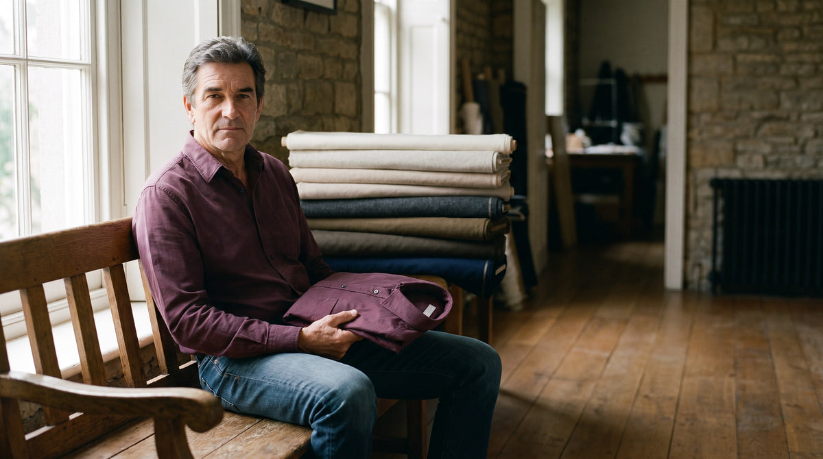 Man seated by a window in a quiet workroom holding a folded shirt