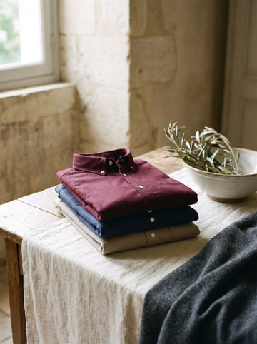 Three folded oxford shirts stacked on cream linen